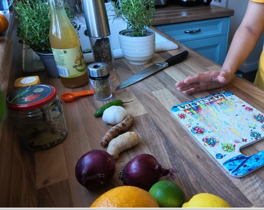 Vista de una tabla de cortar colorida con ingredientes frescos en una mesa de cocina, incluyendo cebollas rojas, jengibre, cúrcuma, limón y lima, así como frascos de especias y una botella de jugo.